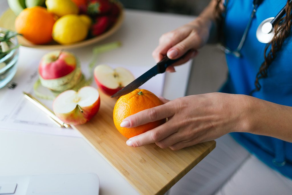 Person's hands slicing an orange and apple on a wooden cutting board in a bright kitchen