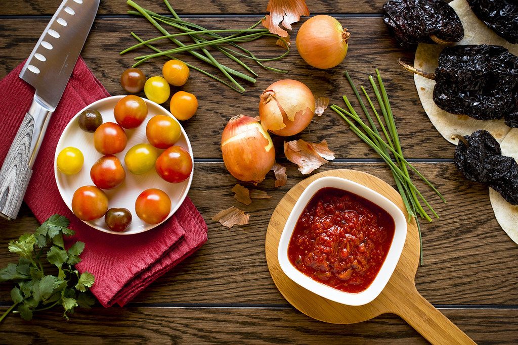 Overhead view of a wooden kitchen counter with cherry tomatoes, onions, chives, herbs, a chef's knife and a bowl of red sauce
