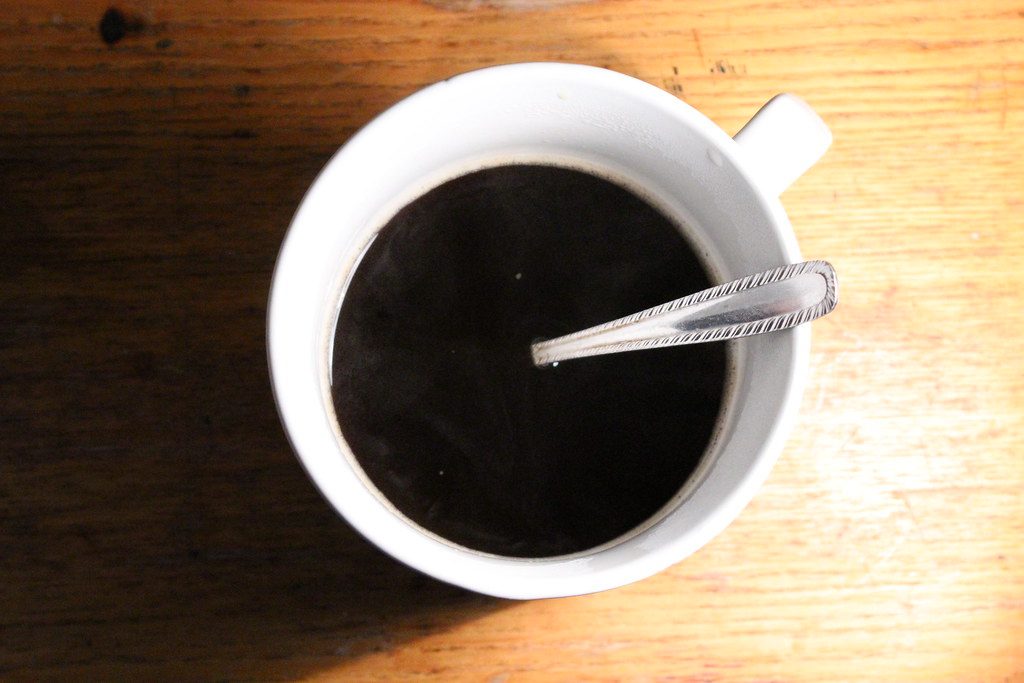 Overhead view of a white cup of dark coffee with a spoon resting on a warm wooden table