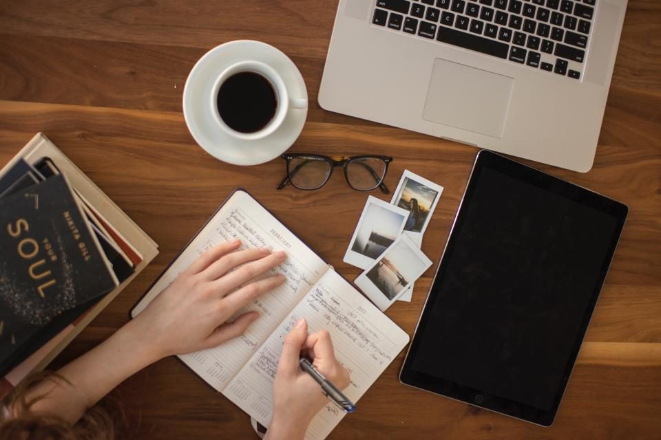 Overhead view of a hand writing in a notebook beside a cup of coffee, laptop, and glasses on a wooden table