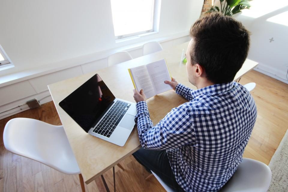Man sitting at a home desk reading a book beside an open laptop by a window