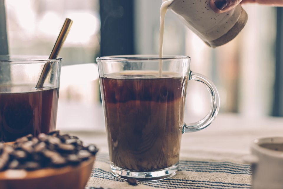 Milk being poured into a glass mug of dark coffee on a table beside a bowl of coffee beans