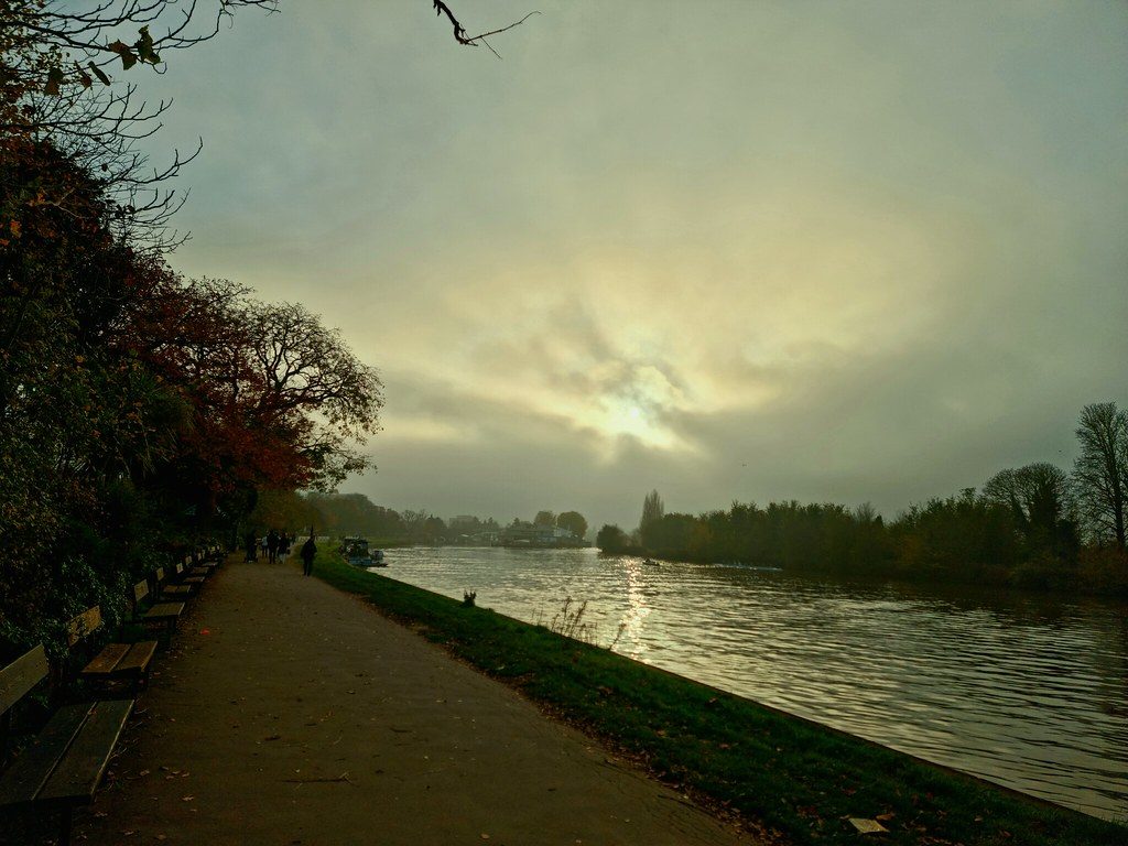 Empty paved path along a calm river at sunrise, with soft golden light through clouds and trees on the left