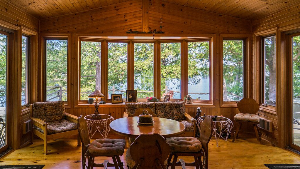 Warm wood-paneled sunroom with a round table and chairs, surrounded by large windows and green trees