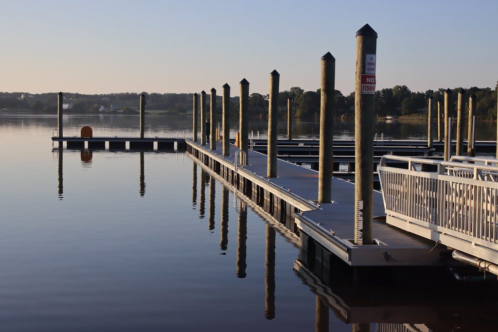 Wooden boat dock with white railing extending into calm lake water at sunrise, reflected in the still surface